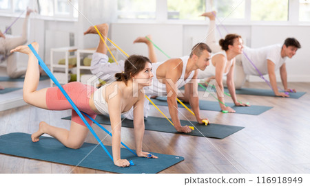Young men and women practicing back leg lift with band standing on knees 116918949