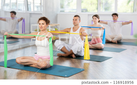 Young men and women doing arm workout with band sitting cross-legged Young men and women doing arm workout with band sitting cross-legged 116918956