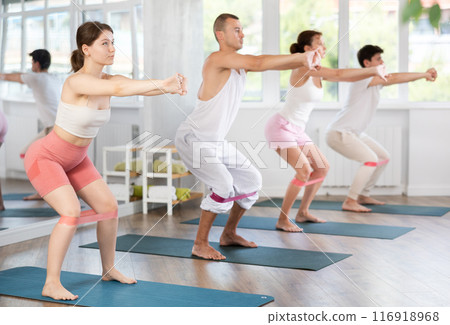 Group of women and men standing and doing stretching exercise with resistance bands in fitness studio 116918968