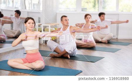 Focused young girl sitting on mat training with resistance band for health wellness, strong biceps or abs muscles during group class in gym health center 116918980
