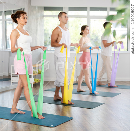 Happy young active woman with group people in sportswear, standing and training exercise with resistance bands in gym 116919007