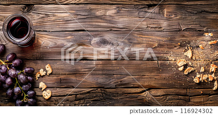 A glass of red wine, a bunch of purple grapes, and scattered bread crumbs rest on a rustic wooden table, offering ample negative space for text or design elements. 116924302