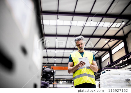 Warehouse employee in warehouse, reading from clipboard. Male worker in reflective clothing in modern industrial factory, heavy industry, manufactrury. 116927162