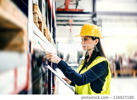 Manager woman in business casual clothes and reflective safety clothes controlling goods in warehouse. 116927180