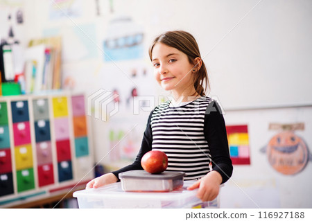 Portrait of schoolgirl, pupil sitting at desk in classroom at the elementary school. Student girl holding apple during snack time, break Portrait of schoolgirl, pupil sitting at desk in classroom at the elementary school. Student girl holding apple during snack time, break 116927188