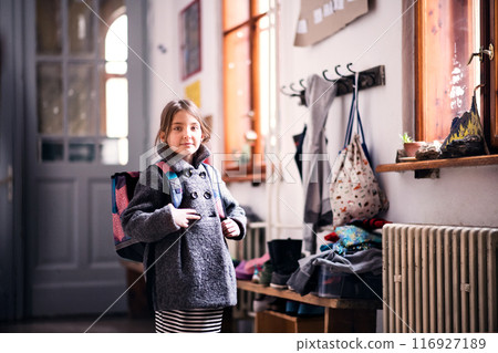 Portrait of schoolgirl, putting coat on by lockers. Student girl with bag in hallway, after day in elementary school. 116927189