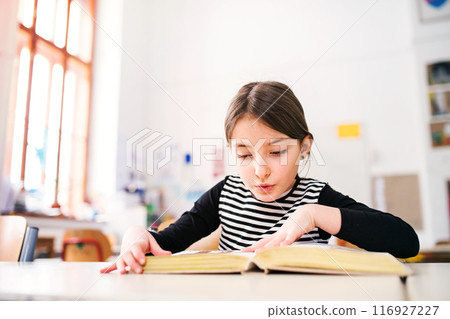 Portrait of cute schoolgirl sitting at desk reading big book. Student girl in classroom in classroom at the elementary school Portrait of cute schoolgirl sitting at desk reading big book. Student girl in classroom in classroom at the elementary school 116927227