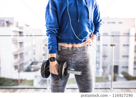 Woman's home workout routine with dumbbell at balcony. Redhead sporty woman working out on terrace with view on city, wearing warm sportswear, close up. Woman's home workout routine with dumbbell at balcony. Redhead sporty woman working out on terrace with view on city, wearing warm sportswear, close up. 116927290