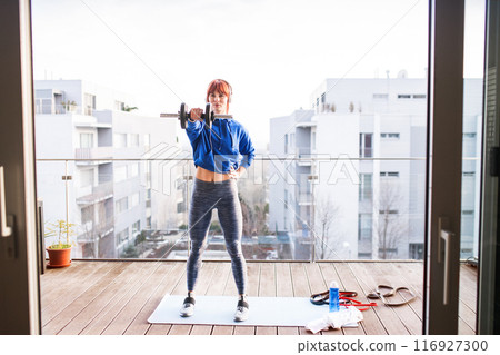Woman's home workout routine with dumbbell at balcony. Redhead sporty woman working out on terrace with view on city, wearing warm sportswear and headphones. 116927300