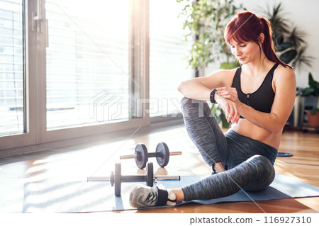 Beautiful redhead woman's home workout routine with dumbbell. Home fitness workout in living room, sporty woman wearing sportswear. Beautiful redhead woman's home workout routine with dumbbell. Home fitness workout in living room, sporty woman wearing sportswear. 116927310