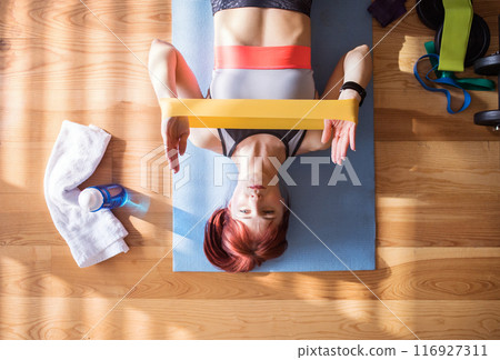 Top view of redhead woman's home fitness routine with resistance band. Home workout on gym mat in living room, sporty woman wearing sportswear. Top view of redhead woman's home fitness routine with resistance band. Home workout on gym mat in living room, sporty woman wearing sportswear. 116927311