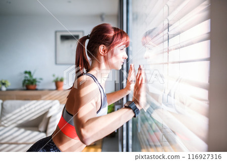Woman's home fitness routine. Beautiful redhead woman working out in living room, standing by window, wearing sportswear. Woman's home fitness routine. Beautiful redhead woman working out in living room, standing by window, wearing sportswear. 116927316