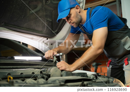The mechanic works with the car engine in the garage. The mechanic uses a wrench to tighten the bolt. The hood of the car is open and the engine is illuminated by a bright light. The mechanic has a The mechanic works with the car engine in the garage. The mechanic uses a wrench to tighten the bolt. The hood of the car is open and the engine is illuminated by a bright light. The mechanic has a 116928690