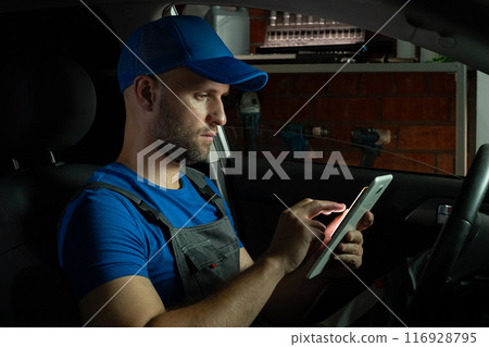 A mechanic sits in the driver's seat of a car in a garage at night, using a tablet computer. 116928795