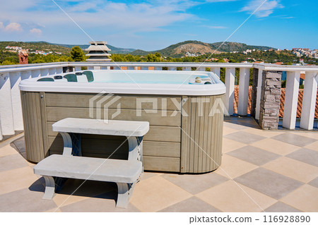 Outdoor hot tub on sunny rooftop deck with bubbling water and comfortable seating with beautiful view on touristic cityscape. Place for relaxation and leisure during summer vacations 116928890