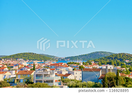 View on coastal town with red roofed houses and lush green hills under clear blue sky. Touristic Mediterranean town near Adriatic sea for summer holidays View on coastal town with red roofed houses and lush green hills under clear blue sky. Touristic Mediterranean town near Adriatic sea for summer holidays 116928895
