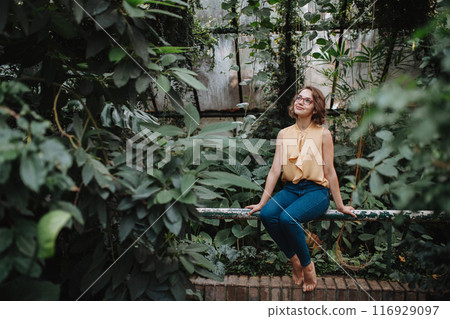 Beautiful woman sitting in botanical garden, in the middle of lush green foliage, looking at camera. 116929097