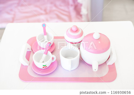 A set of pink white pastel color wood  toy tea cups including 2 coffee cups with teaspoons, a teapot, a sugar jar  and a milk jug is placed on a tray. 116930656