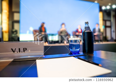 VIP seating, with plain white paper, a pencil, a glass of water, and a bottle of water on a desk with a blurred background in the seminar room. VIP seating, with plain white paper, a pencil, a glass of water, and a bottle of water on a desk with a blurred background in the seminar room. 116930962