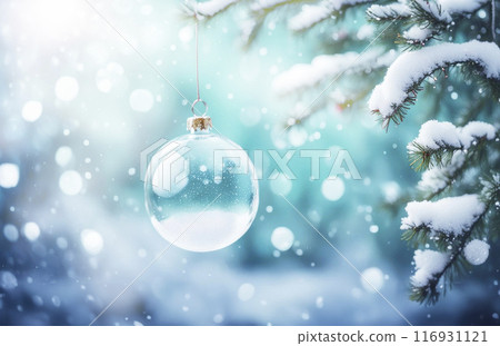 A transparent white glass New Year's ball hangs on a red ribbon on a green branch covered with snow. Light blue background with bokeh effect and snowflakes in blurred focus 116931121
