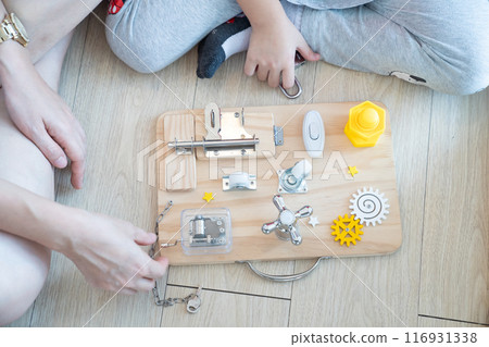 Child and mother are playing with busy board toy on the wooden floor together. Children educational busy board toy for developing motor skills. 116931338