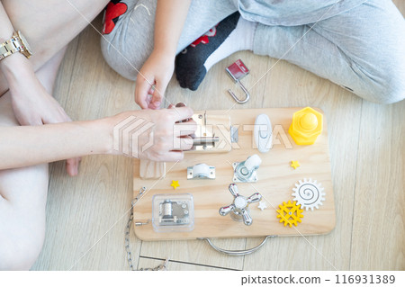 Child and mother are playing with busy board toy on the wooden floor together. Children educational busy board toy for developing motor skills. 116931389