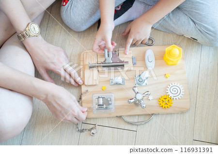 Child and mother are playing with busy board toy on the wooden floor together. Children educational busy board toy for developing motor skills. Child and mother are playing with busy board toy on the wooden floor together. Children educational busy board toy for developing motor skills. 116931391