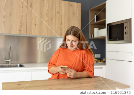 Portrait of smiling young housewife using phone in modern kitchen. Home concept. Copy space. 30s Woman watching recipe on social media, video tutorial how to prepare meal. 116931873