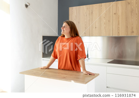 Portrait of smiling young housewife standing near counter in modern kitchen. Home concept. Copy space. 30s Woman dreaming at new home 116931874