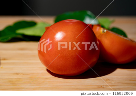 Tomato and sliced half of tomato beside it, on wood plate in studio light with dark theme. Tomato and sliced half of tomato beside it, on wood plate in studio light with dark theme. 116932009