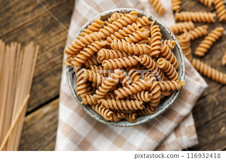 Uncooked whole grain pasta. Raw fusilli pasta in bowl on wooden table. Top view. 116932418