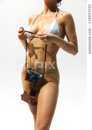 Cropped image of attractive woman with tanned slim body, wearing white swimsuit and holding vintage camera against light background 116933293