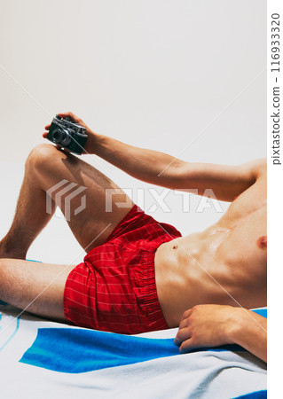 Cropped image of tanned, muscular male body, torso, model in red swim shorts lying on beach towel with vintage camera 116933320