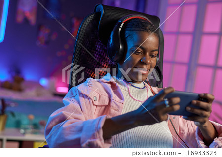 Portrait of teenage African American girl playing mobile games while relaxing at home at night 116933323