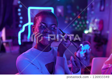 Portrait of smiling young Black woman testing luminescent makeup in dark room with neon lights, copy space Portrait of smiling young Black woman testing luminescent makeup in dark room with neon lights, copy space 116933349