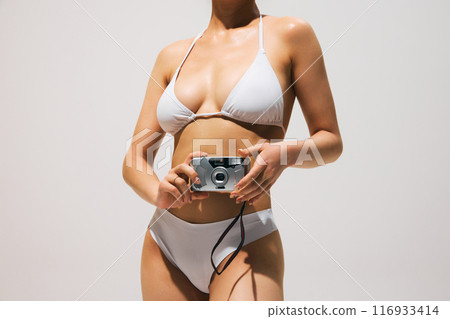 Cropped image of young women with slim body, posing in white swimsuit with retro photo camera against light grey background. 116933414
