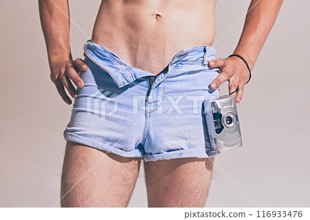 Cropped image of shirtless muscular man, wearing denim shorts and standing with vintage camera, creating nostalgic vibes 116933476