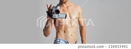 Retro Tech. Cropped image of shirtless man with muscular, athletic body, in denim short standing with vintage camcorder and filming 116933491
