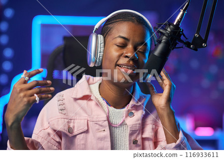 Portrait of inspired African American girl singing to microphone and recording music in home studio with pink neon lights copy space 116933611