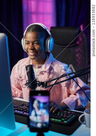 Vertical portrait of young African American woman wearing headphones and playing videogames with online stream Vertical portrait of young African American woman wearing headphones and playing videogames with online stream 116933682