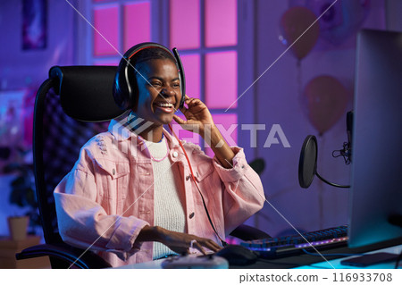 Portrait of smiling African American young woman playing video games at night and recording online stream with neon lighting 116933708