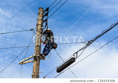 A man performing maintenance work on a utility pole with a blue sky in the background 116934306
