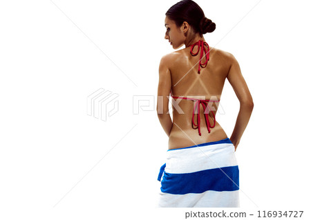 Woman holds towel against her buttocks, her glistening, deeply tanned skin showing clear tan lines against white studio background. 116934727