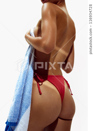 Woman wraps herself in towel, revealing tan marks, lines of bikini on her deeply tanned skin against white studio background. 116934728