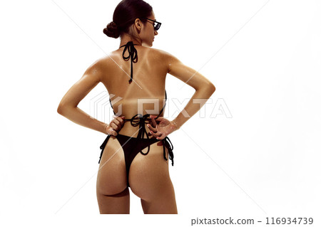 Woman, with shimmering skin, adjusts her bikini bottoms, highlighting pronounced tan lines on her deeply bronzed body against white studio background. 116934739