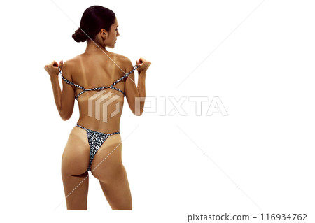 Woman adjusts bikini, showing distinct tan lines on her deeply tanned skin, indicating she sunbathing extensively against white studio background. 116934762