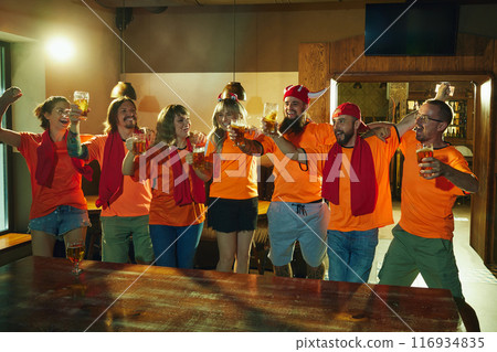 Friends, all in orange shirts and red scarves, cheerfully clink glasses, singing of joy and happiness, celebrating win of favorite team at bar. Friends, all in orange shirts and red scarves, cheerfully clink glasses, singing of joy and happiness, celebrating win of favorite team at bar. 116934835