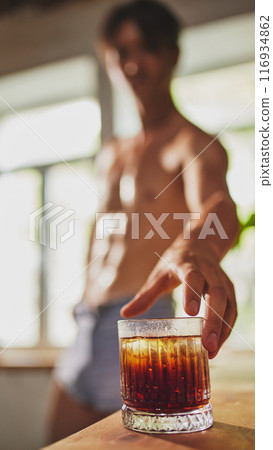 Shirtless man in denim shorts reaches for glass of iced dark beverage on table, with blurred background of sunlit room. Shirtless man in denim shorts reaches for glass of iced dark beverage on table, with blurred background of sunlit room. 116934862