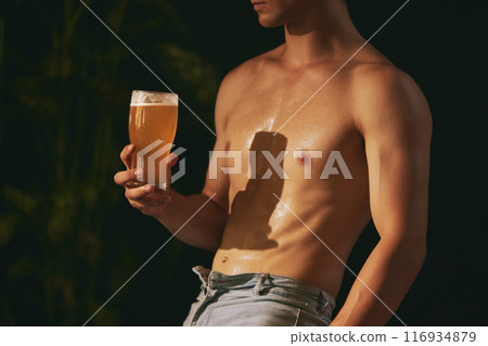 Shirtless man with perfect, athletic body leaned on wooden bar counter and drinks cold, refreshing, foamy beer. 116934879