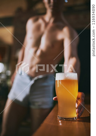 Fit, shirtless man in denim shorts leans towards tall, frothy craft glass of beer on wooden bar. Relaxed atmosphere. Oktoberfest. 116934890